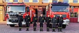 Strażacy w ubraniach koszarowych stoją przed samochodami strażackimi. W tle budynek straży pożarnej.