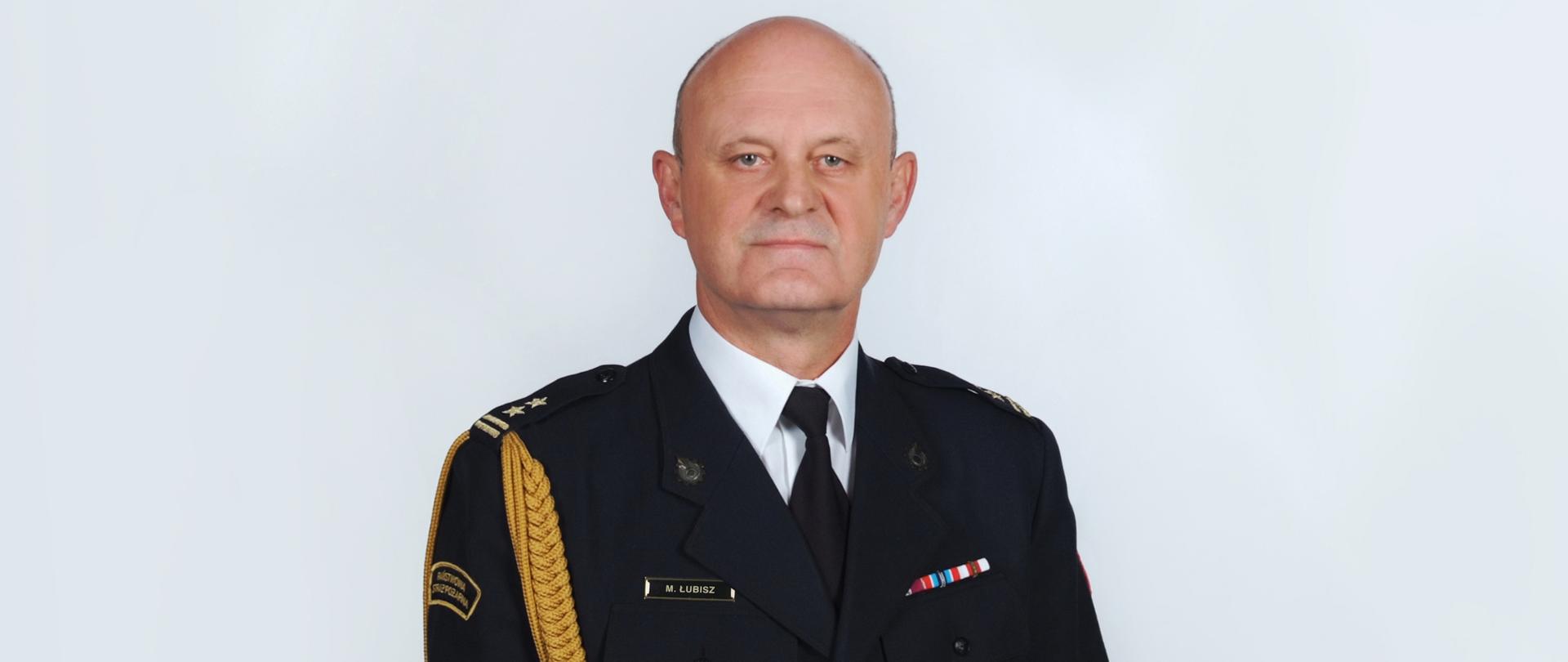 Na zdjęciu brygadier Maciej Łubisz, Zastępca Komendanta Powiatowego Państwowej Straży Pożarnej w Pabianicach. Występuje w umundurowaniu wyjściowym ze sznurem. Na lewej piersi przypięte baretki. Na prawej piersi od góry znajduje się imiennik oraz absolwentka Szkoły Głównej Służby Pożarniczej.