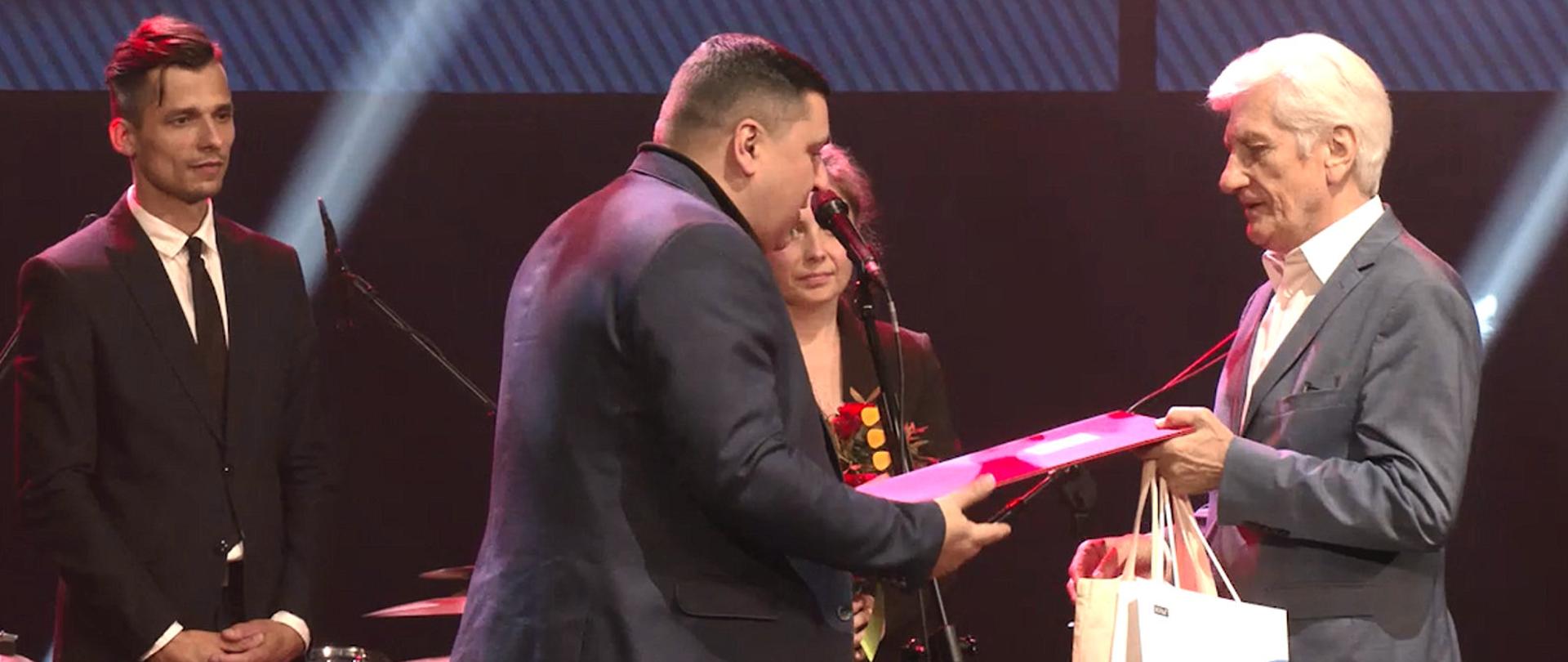 Zdjęcie w poziomie. Na zdjęciu cztery osoby ujęte w półpostaci. Minister Andrzej Sabatowski wręcza laureatowi dyplom