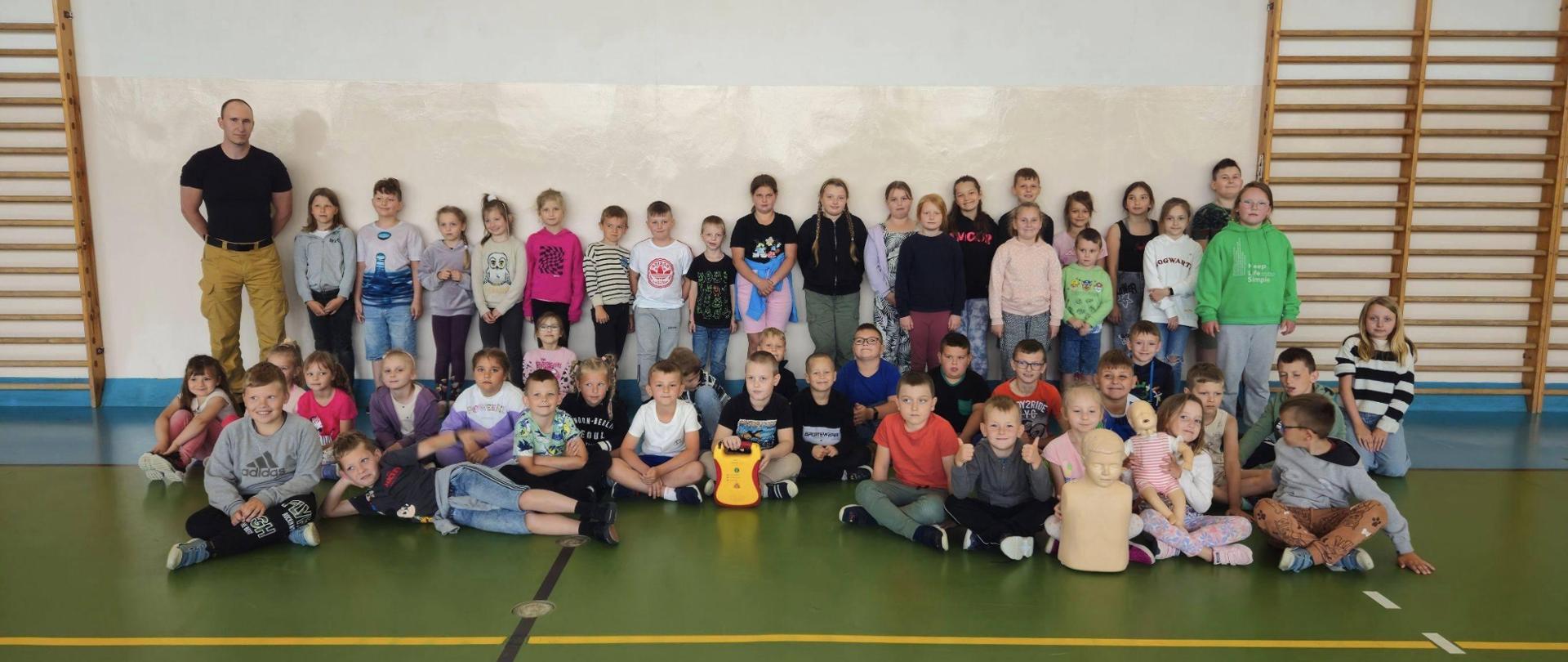 Sala gimnastyczna. Pod ścianą przy drabinkach stoi grupa dzieci ze strażakiem.