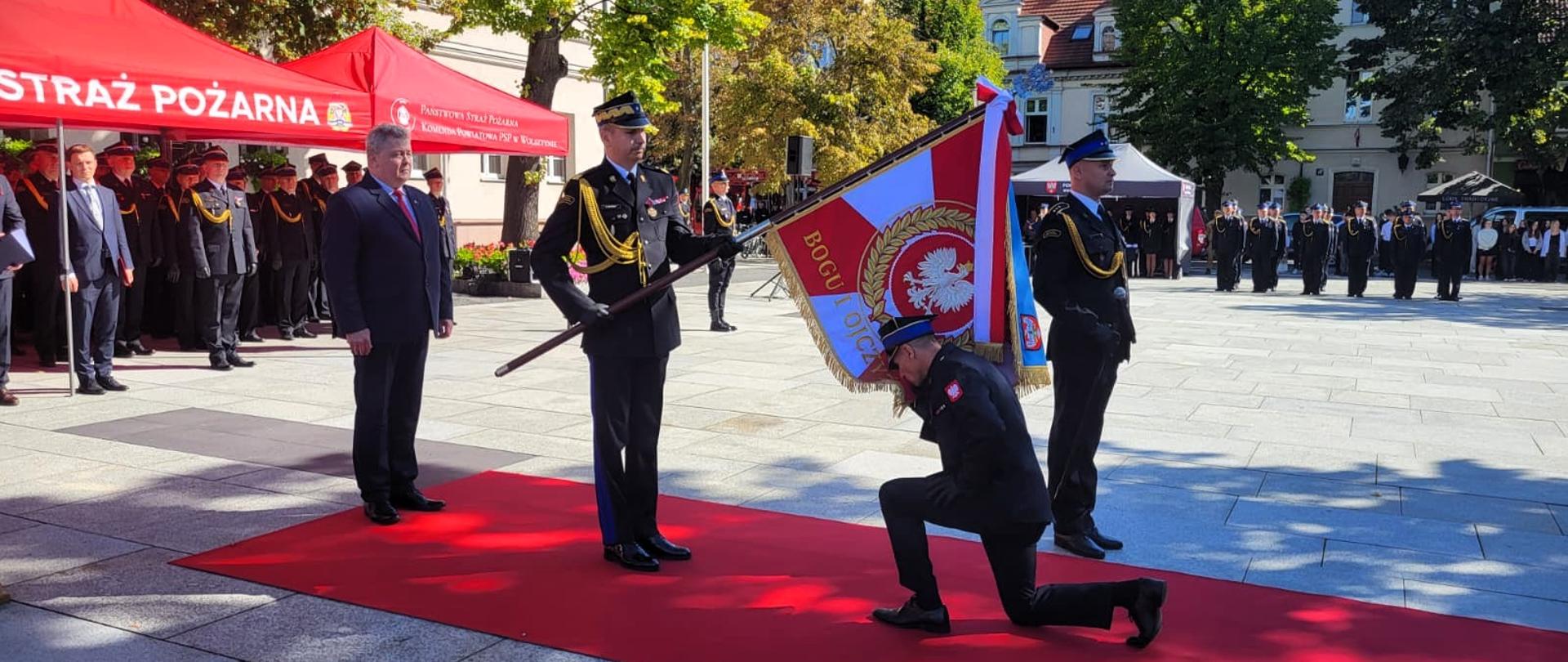 Uroczysty moment oddania honorów sztandarowi PSP – strażak w mundurze galowym klęczy i całuje sztandar z napisem „Bogu i Ojczyźnie”; obok funkcjonariusz trzymający drzewiec, w tle oficjele i czerwona strefa z namiotami PSP.