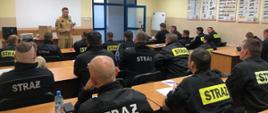 Spotkanie organizacyjne w Ośrodku Szkolenia Komendy Wojewódzkiej PSP w Krakowie. Widok z tyłu. Słuchacze szkolenia w ławkach. Prowadzący na środku sali, przekazuje informacje organizacyjne. 
