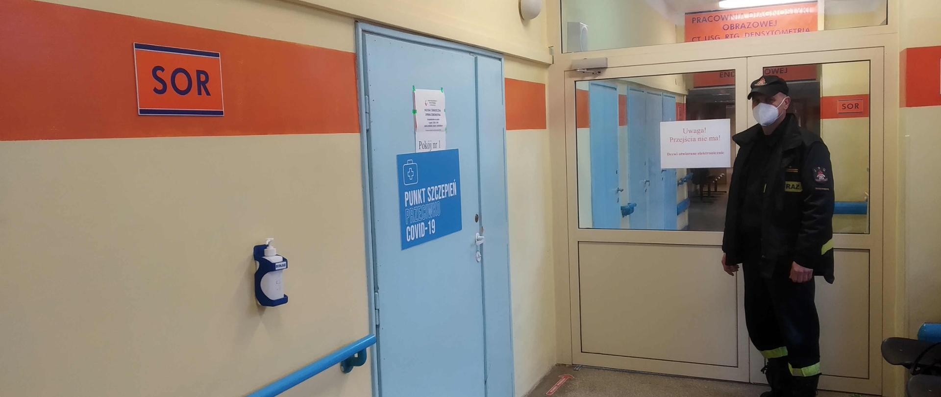 Zdjęcie zrobione na szpitalnym korytarzu. Po lewej stronie znajdują się niebieski drzwi z napisem punkt szczepień przeciwko covid-19. Naprzeciwko drzwi stoi strażak ubrany w ubranie koszarowe, który oczekuje na szczepienie. Twarz ma zasłoniętą maseczką. Za nim znajdują się oszklone drzwi prowadzące do dalszej części szpitala.