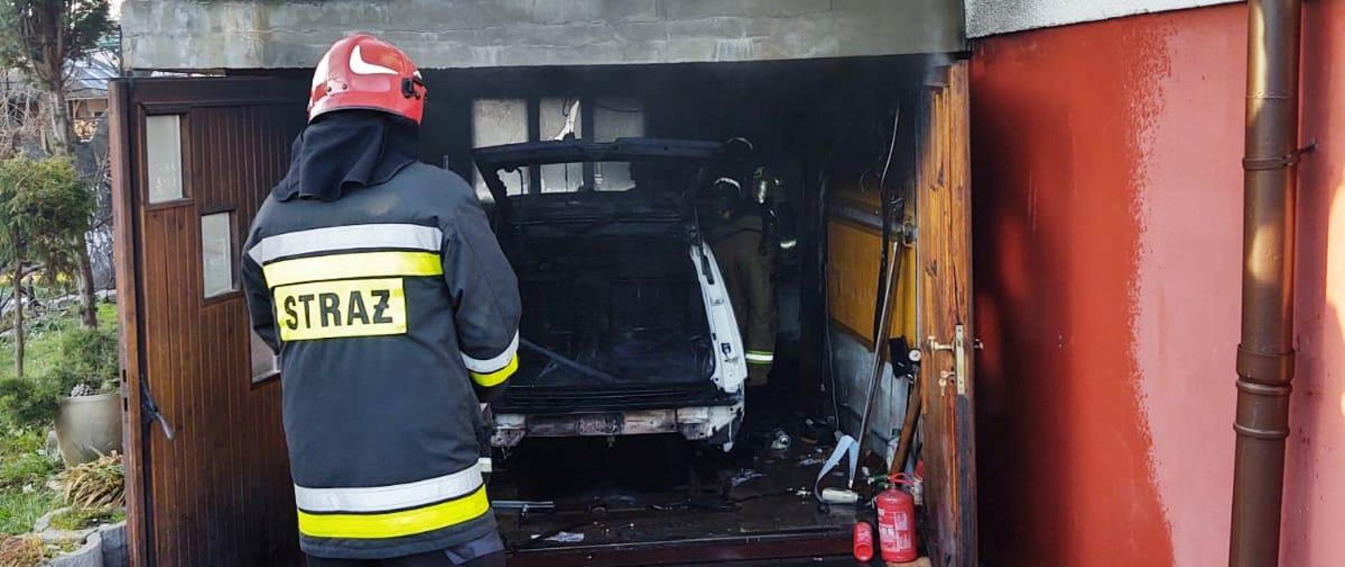 Widok garażu zlokalizowanego przy budynku mieszkalnym a w nim spalony samochód. Przed garażem stoi strażak uczestniczący w działaniach.