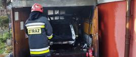 Widok spalonego samochodu znajdującego się w garażu zlokalizowanym przy budynku jednorodzinnym a przed garażem stojący strażak. 