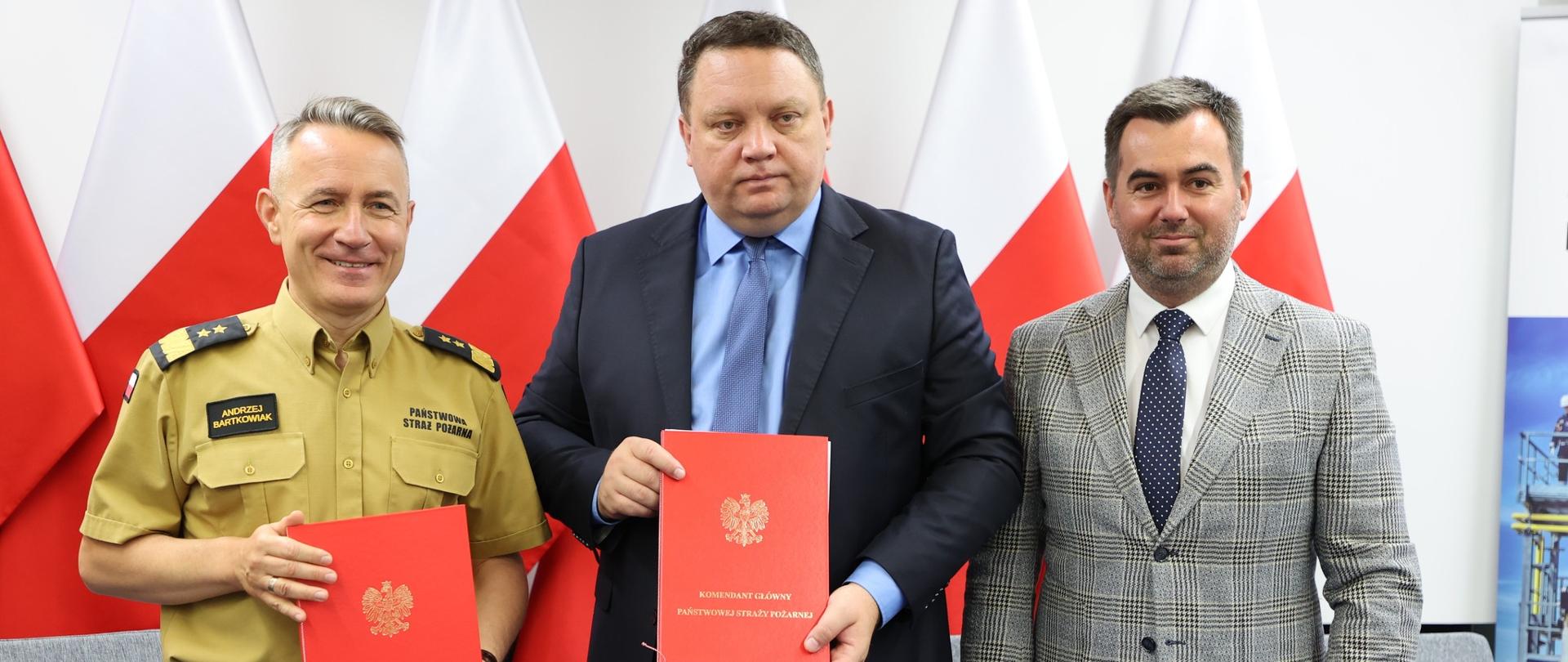 Trzej mężczyźni. Dwaj w garniturach, jeden w piaskowym umundurowaniu, trzymający czerwone teczki. W tle flagi narodowe