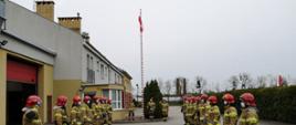 Strażacy stoją w dwóch rzędach na przeciwko siebie, uroczyście podnosząc flagę Rzeczypospolitej Polskiej na maszt.