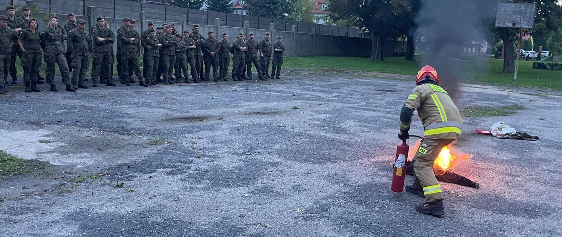 Trwa pokaz. Strażak gasi kontrolowalny pożar gaśnicą . Z lewej strony stoi i obserwuje to grupa żołnierzy.