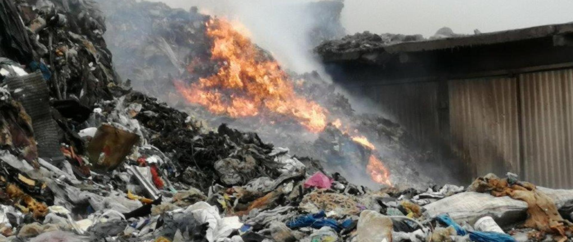 Zdjęcie obrazuje usypaną pryzmę śmieci, w tle widać jak część z nich się pali jasno-żółtym i pomarańczowym płomieniem. Widoczne odpady to tworzywa sztuczne, torby foliowe, butelki plastykowe itp. W prawej części zdjęcia widać jak płomienie z palących się odpadów powoli obejmują ściany budynku garażowego. W górnej części zdjęcia widać duże jasne zadymienie.