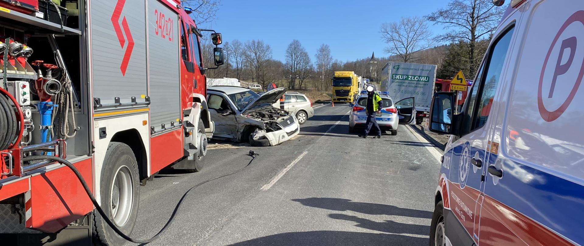 Na zdjęciu widać czerwony samochód strażacki, samochód ma otwartą jedną skrytkę w której widać asortyment do działań strażackich. Przed samochodem znajduje się biały pojazd z uszkodzonym zderzakiem i podniesioną maską. Samochód jest koloru białego. W oddali widać żółtą ciężarówkę. Po prawej stronie zdjęcia widać radiowóz strażacki obok, którego stoi funkcjonariusz policji. Po prawej stronie zdjęcia widać połowę karetki pogotowia. 