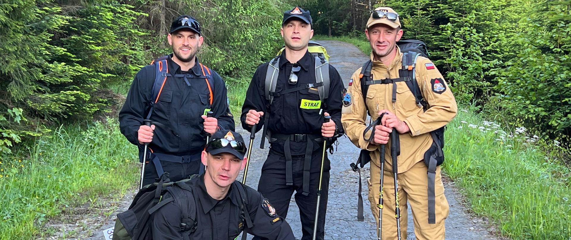 Zdjęcie przedstawia trzech strażaków ubranych w umundurowanie koszarowe oraz jednego strażaka w umundurowaniu służbowym. Strażacy mają na plecach plecaki oraz trzymają w rękach kijki do maszerowania. W tle widoczny jest las.