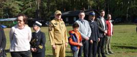 Pomorski komendant wojewódzki Państwowej Straży Pożarnej stoi wśród zaproszonych gości na tle lasu.