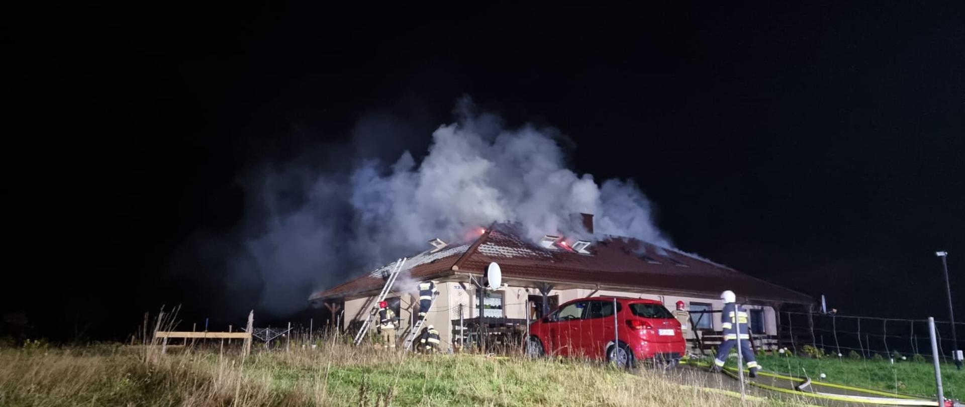 Na zdjęciu w porze nocnej widać budynek z widocznym dymem wydobywającym się z dachu. Obok stoi czerwony samochód na podjeździe do garażu. Przy budynku stoją strażacy gaszący pożar z zewnątrz.