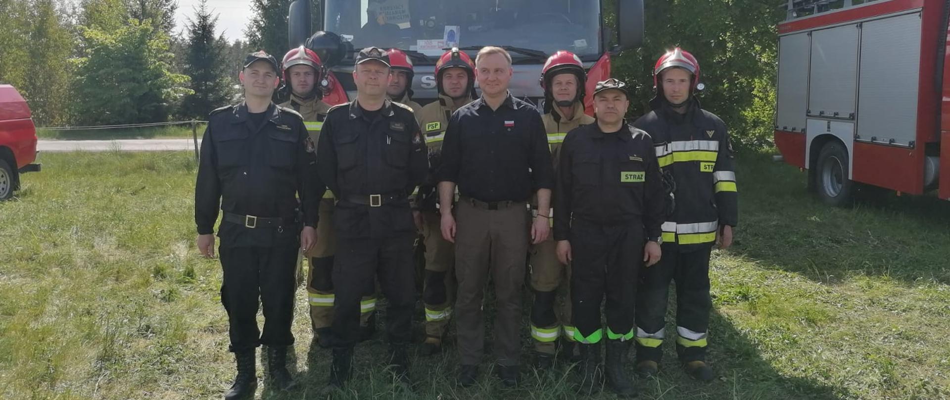Strażacy zabezpieczający lądowisko rządowego śmigłowca stoją wraz z prezydentem Andrzejem Dudą przed wozem strażackim.