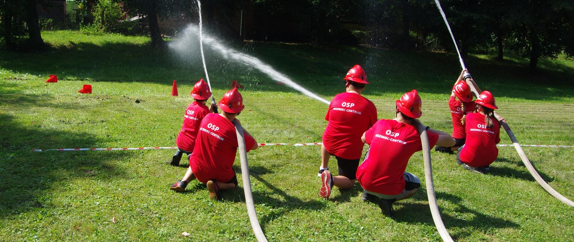 Widok z tyłu. Członkowie MDP w czerwonych koszulkach i czerwonych hełmach strumieniami wody strącają pachołki w konkurencji „mini-ćwiczenia bojowego”.