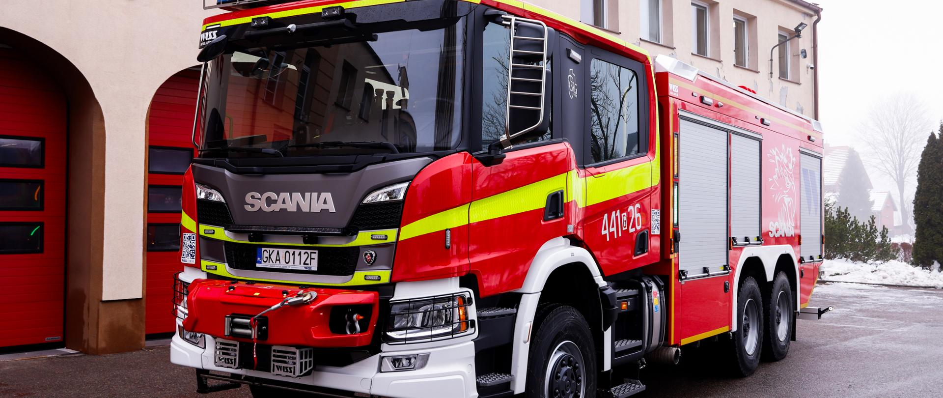 Zdjęcie przedstawia nowy ciężki samochód ratowniczy na podwoziu marki Scania. W tle widać budynek kartuskiej straży pożarnej