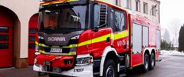 Zdjęcie przedstawia nowy ciężki samochód ratowniczy na podwoziu marki Scania. W tle widać budynek kartuskiej straży pożarnej