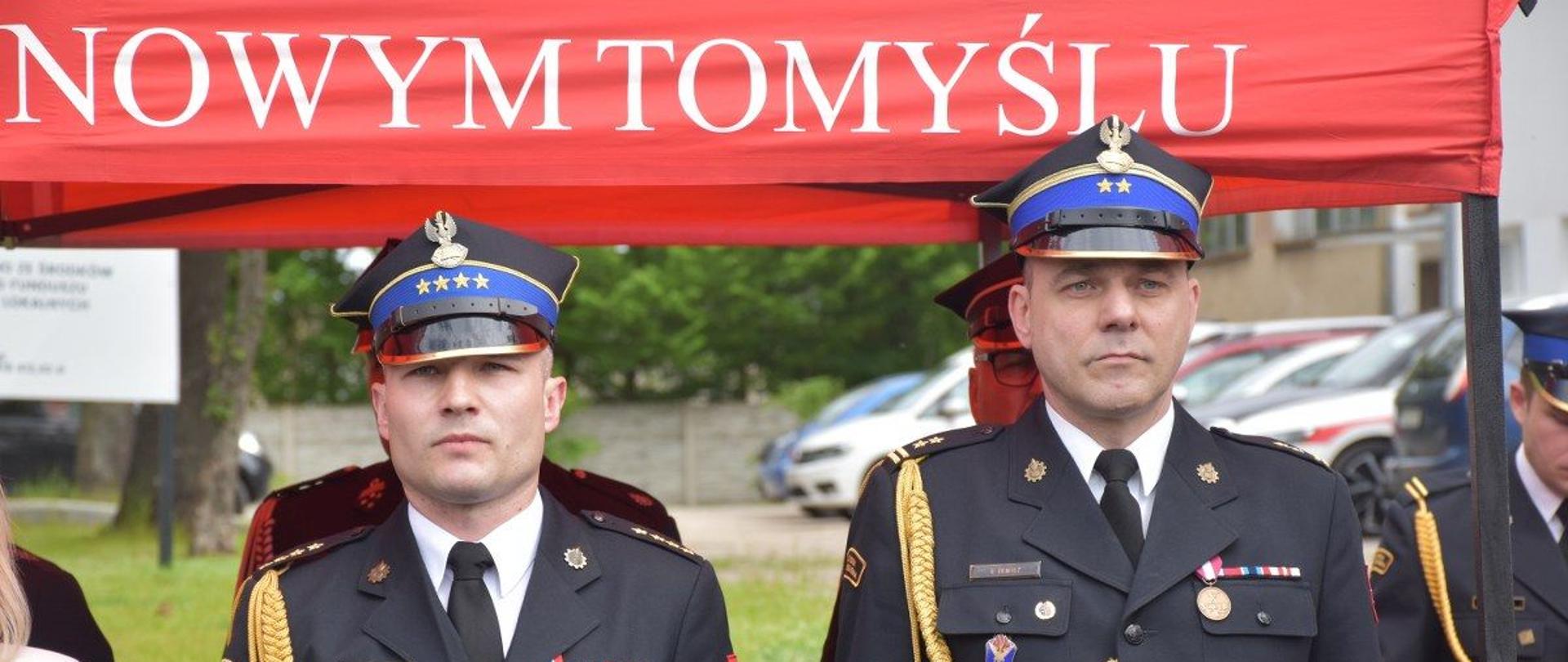 dwóch strażaków q mundurach ze sznurem i w rogatywkach stoi pod czerwonym namiotem