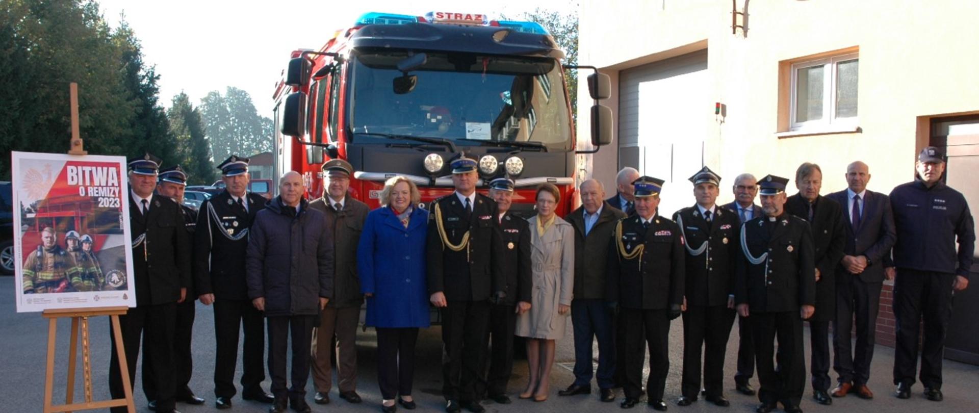 a asfaltowym placu stoi samochód pożarniczy (widok z przodu). Przed nim stoi kilkanaście osób, w tym strażacy w mundurach PSP i OSP oraz osoby cywilne, w tym 2 kobiety. Po lewej stronie na stojaku plakat akcji bitwa o remizy 2023. Po prawej stronie zdjęcia fragment budynku Komendy z bramą garażową, po lewej zaparkowane samochody osobowe, dalej drzewa.