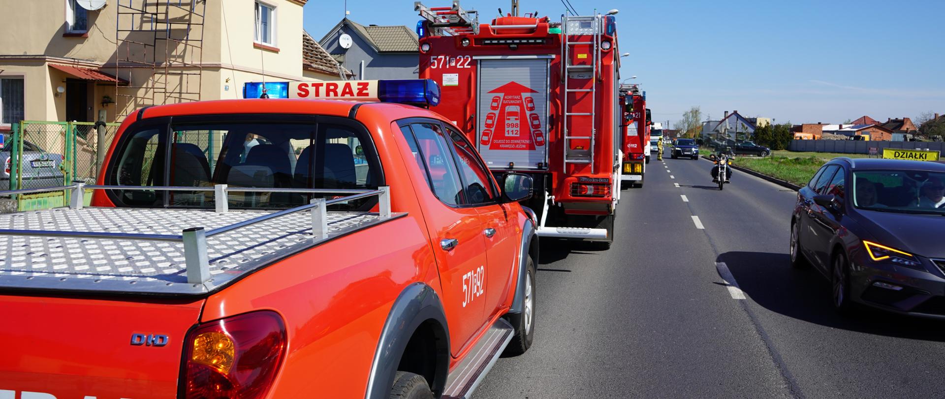 Z lewej strony na drodze stoją wozy strażackie. Z prawej strony jadą auta osobowe i motocyklista.
