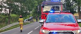 Strażacy zabezpieczają miejsce zdarzenia przez odpowiednie ustawienie samochodów pożarniczych na drodze w miejscu wypadku.