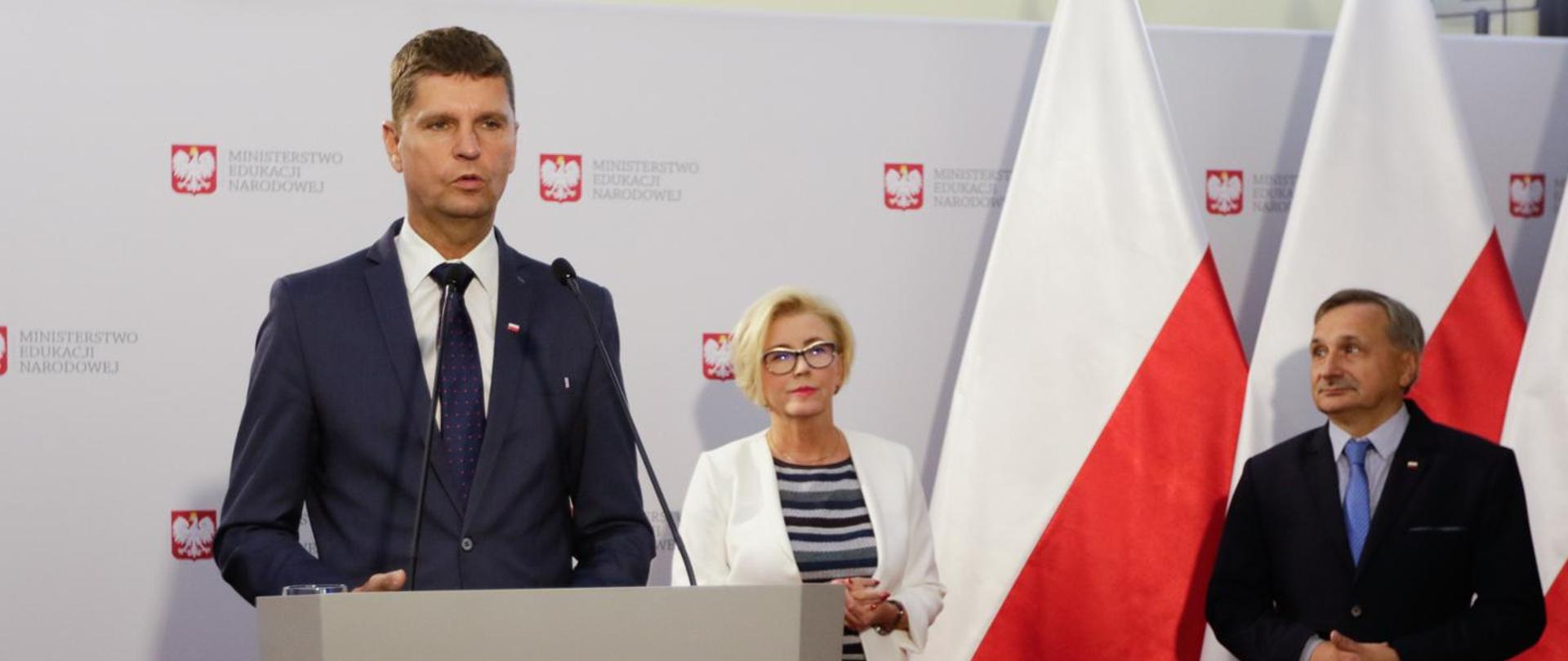 Na pierwszym planie widać elegancko ubranego Ministra Edukacji Narodowej Dariusza Piontkowskiego, który przemawia podczas konferencji prasowej. Na drugim planie widać kobietę - Sekretarza Stanu Marzenę Machałek oraz mężczyznę - Podsekretarza Stanu Macieja Kopcia na tle biało czerwonej flagi.