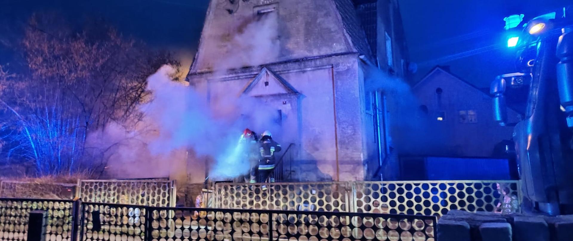 Widoczni strażacy-kierowcy przed budynkiem, w którym ma miejsce pożar piwnicy