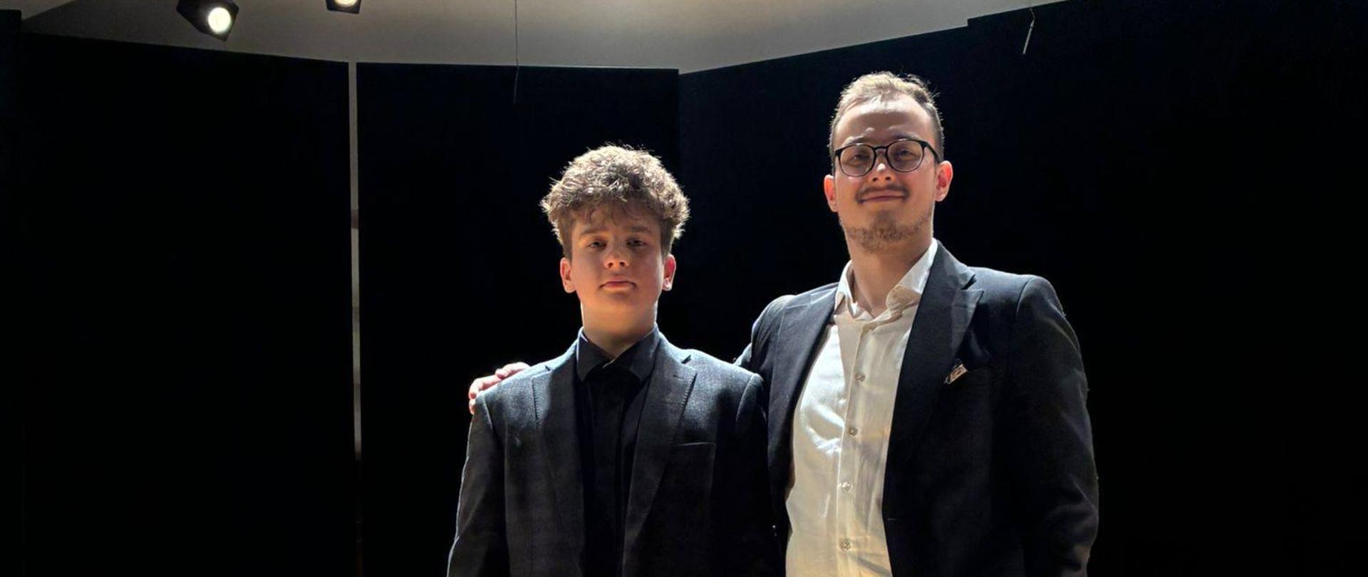 Zdjęcie pamiątkowe dwóch mężczyzn stojących na środku oświetlonej sceny na tle czarnej horyzontu. Po lewej młody chłopak w ciemnym garniturze i czarnej koszuli, po prawej nieco starszy mężczyzna w okularach, ciemnej marynarce i białej rozpiętej koszuli, trzymający rękę na ramieniu towarzysza.