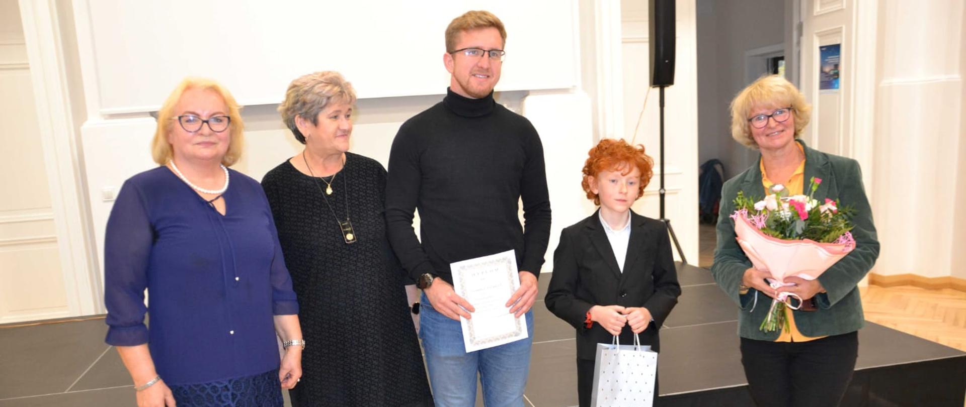 Stojące osoby pozujące do zdjęcia od prawej Pani Liliana Stopka trzymająca kwiaty, uczeń Gabryś Charęza trzymający upominki, nauczyciel Tomasz Brusiński trzymający dyplom i obok dwie stojące panie.