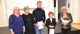 Stojące osoby pozujące do zdjęcia od prawej Pani Liliana Stopka trzymająca kwiaty, uczeń Gabryś Charęza trzymający upominki, nauczyciel Tomasz Brusiński trzymający dyplom i obok dwie stojące panie.