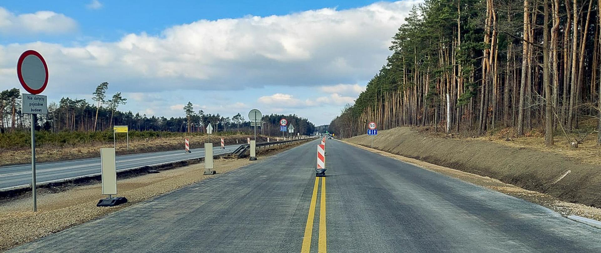 Fotografia przedstawia drogę w trakcie budowy lub remontu. Na pierwszym planie widoczny jest znak zakazu wjazdu z napisem "Nie dotyczy pojazdów budowy". Droga jest oznaczona żółtymi liniami, a na środku jezdni ustawione są pachołki ostrzegawcze. Po prawej stronie drogi znajduje się las, a po lewej stronie widać inne znaki drogowe oraz barierki ochronne. Na dalszym planie widoczne są znaki ograniczenia prędkości do 50 km/h oraz znak informujący o zwężeniu jezdni.