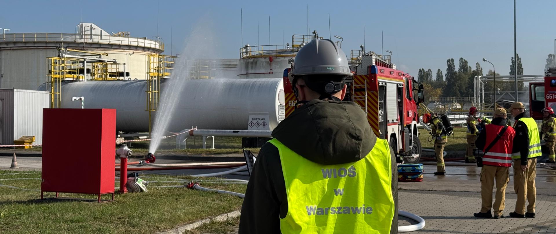 Na pierwszym planie stoi Inspektor Wojewódzkiego Inspektoratu Ochrony Środowiska w Warszawie, który obserwuje i dokumentuje przebieg ćwiczeń. W tle, po lewej stronie, znajdują się duże, białe zbiorniki przemysłowe. Z jednego z hydrantów wodnych tryska strumień wody, skierowany w stronę tych zbiorników. Po prawej stronie znajduje się wóz strażacki, a wokół niego kilku strażaków w pełnym umundurowaniu ochronnym prowadzi działania. Całość odbywa się na placu z kostki brukowej, obok trawiastego terenu. Niebo jest bezchmurne, a warunki pogodowe słoneczne.