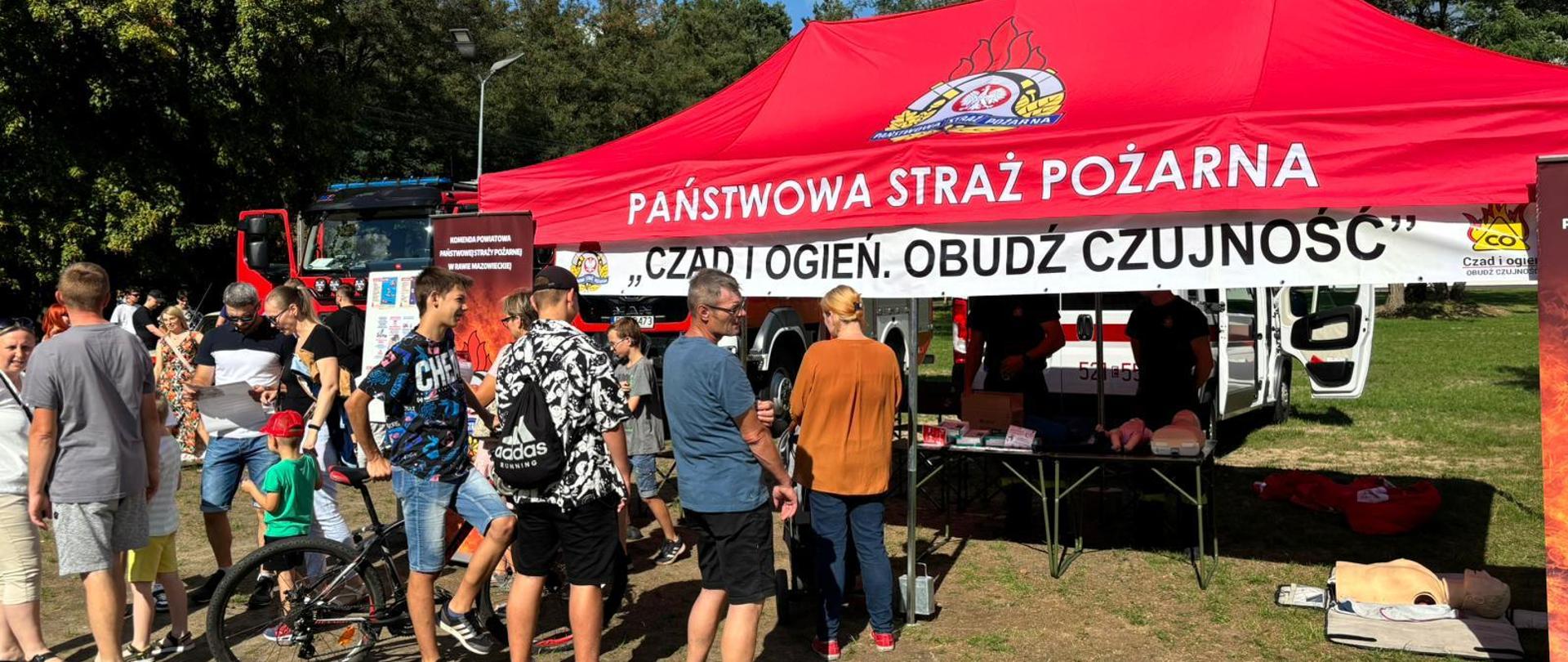 Na zdjęciu widać czerwony namiot z logiem PSP oraz napisem Państwa Straż Pożarna "Czad i ogień. Obudź czujność", a przed nim stoi kilkoro ludzi.