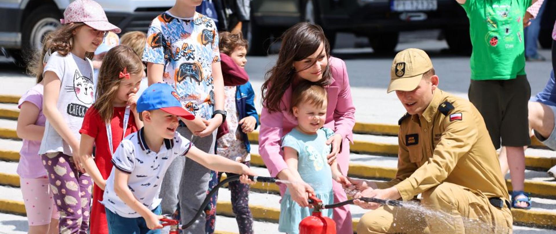 kobieta w różowym stroju wraz z małą dziewczynką trzymają gaśnicę, pomaga im strażak, za nimi dzieci czekają w kolejce, obok kobiety stoi dziecko z gaśnicą
