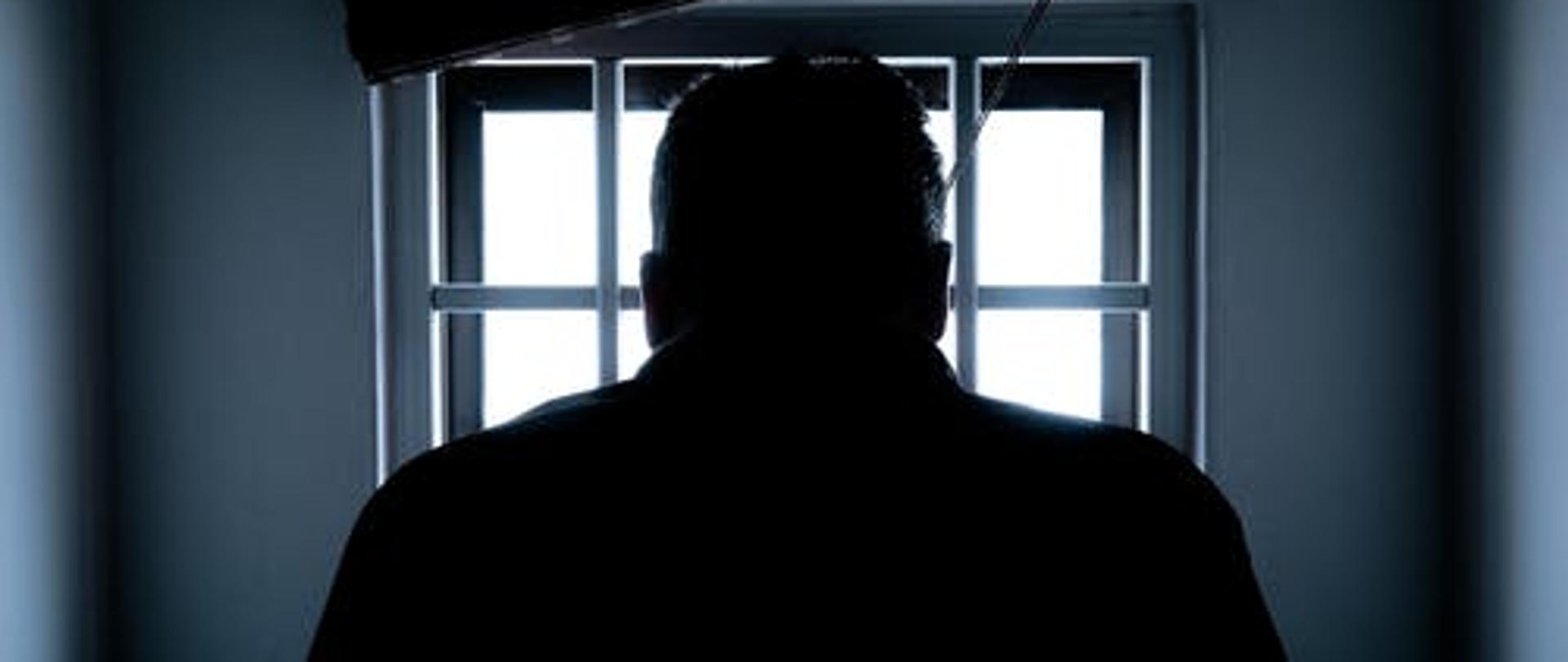 Cela. Okno zakratowane. W środku odwrócony plecami człowiek.
