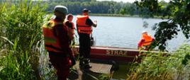 Na zdjęciu strażacy przygotowujący się do ćwiczę na akwenie wodnym. widoczna jest łódź ratownicza ze strażakiem w środku, oraz strażacy ubrani w kamizelki wypornościowe , którzy za chwilę wejdą do łodzi, żeby realizować zamierzone ćwiczenie.