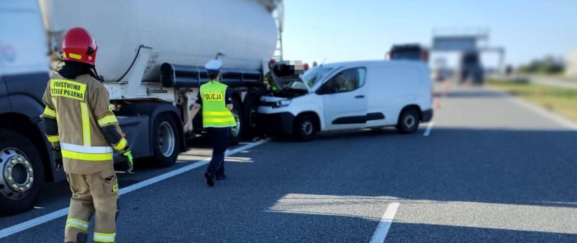 Na zdjęciu widoczne jest miejsce zdarzenia drogowego. W prawym pasie ruchu znajduje się biały samochód dostawczy, którego przód uderzył w tylną część naczepy samochodu ciężarowego typu cysterna. Przód pojazdu dostawczego jest zgnieciony. Na miejscu obecni są: funkcjonariusz Policji oraz strażak Państwowej Straży Pożarnej, którzy prowadzą działania zabezpieczające miejsce zdarzenia. W tle widoczne są ustawione pachołki drogowe.