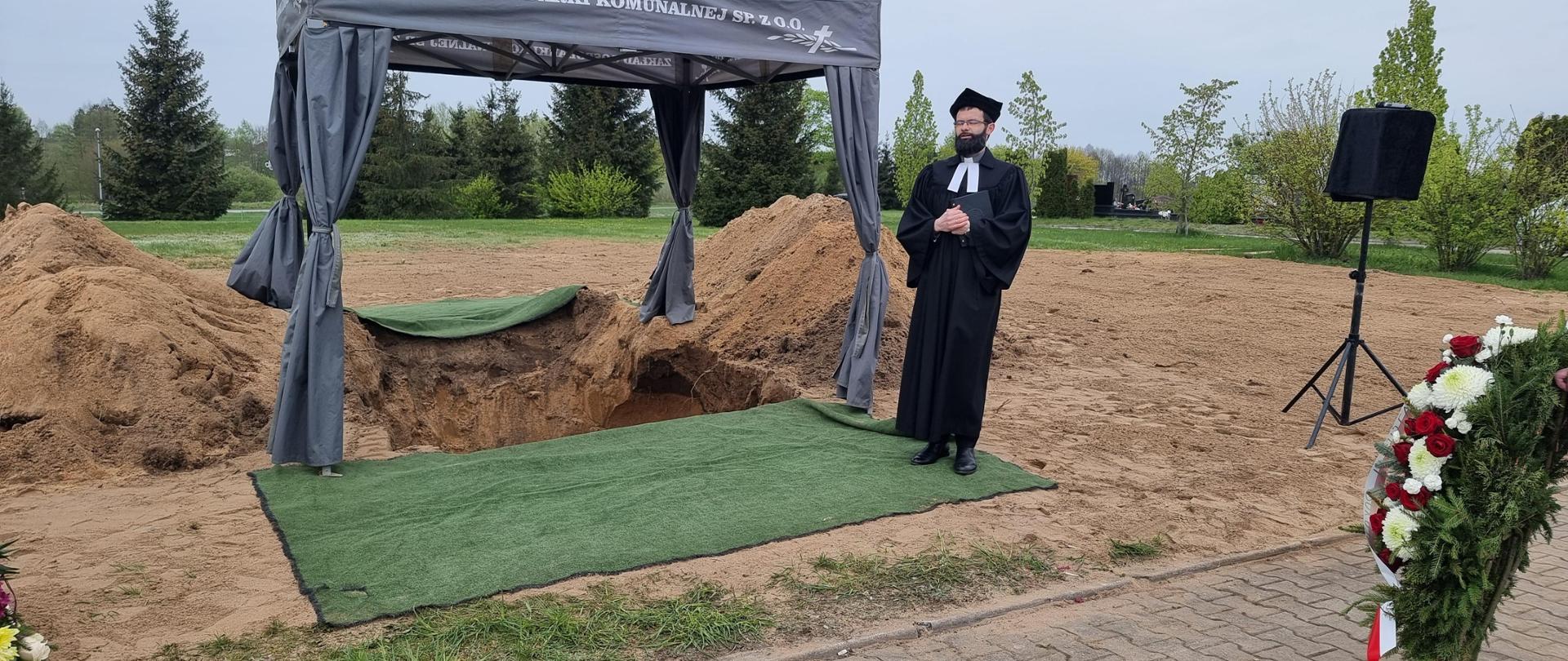 Namiot pogrzebowy, pod którym jest wykopany grób, przy namiocie stoi ksiądz. Obok stoi wieniec oraz sprzęt nagłaśniający.