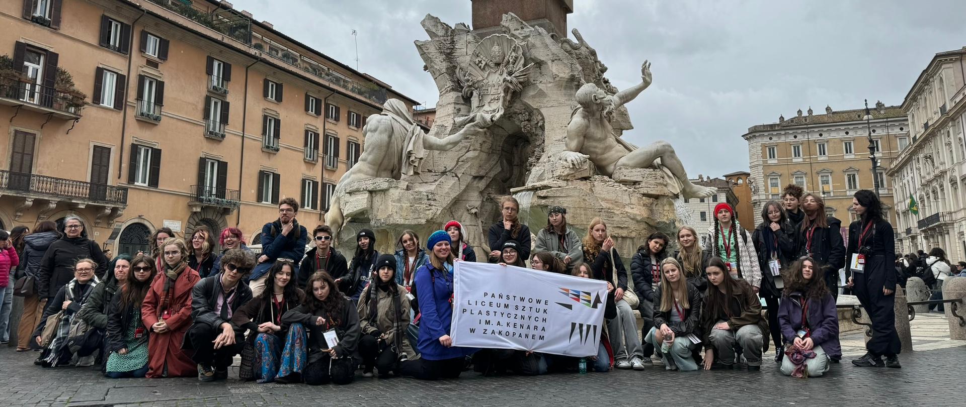 Rzym, Piazza Navona, Fontanna Czterech Rzek, zdjęcie grupowe z banerem PLSP im. A. Kenara, uczestnicy wycieczki: od lewej w dolnym rzędzie – mgr Ewa Ross-Baczyńska, mgr Katarzyna Skórka-Trepka, Joanna Pitoń kl. 2c, Natasza Cisoń kl. 2c, Mateusz Walkosz kl. 2b, Klaudia Biedrawa kl. 2b, Helena Bryja kl. 2b, Julia Kukawska kl. 3a, Kornelia Furca kl. 5a, Zuzanna Mitana kl. 2c, Wiktoria Zwijacz kl. 2c, Oliwia Szatanek kl. 2c, Martyna Plewa kl. 2c, Natalia Bienias kl. 2c, Amelia Kutwin kl. 2b; w górnym rzędzie – s. Grażyna Powierża, Anna Remiasz, Zuzanna Pietrzyk, Gabriela Głód, Mateusz Baczyński, Mateusz Mateusiak kl. 2a, Eve Gardner kl.2a, Maria Kolasa kl. 2c, Paulina Bobak kl. 2b, Tymoteusz Kardaś kl. 2b, Krystyna Skrobot kl. 2c, Martyna Laszczyk kl. 2c, Hanna Bodziony kl. 2a, Marcelina Bobak kl. 2c, Zuzanna Trzop kl. 2c, Klaudia Tyrała kl. 2b, Anna Olejarz kl. 3a, Natalia Łuszczek kl. 2b, Maja Sochacka kl. 3a