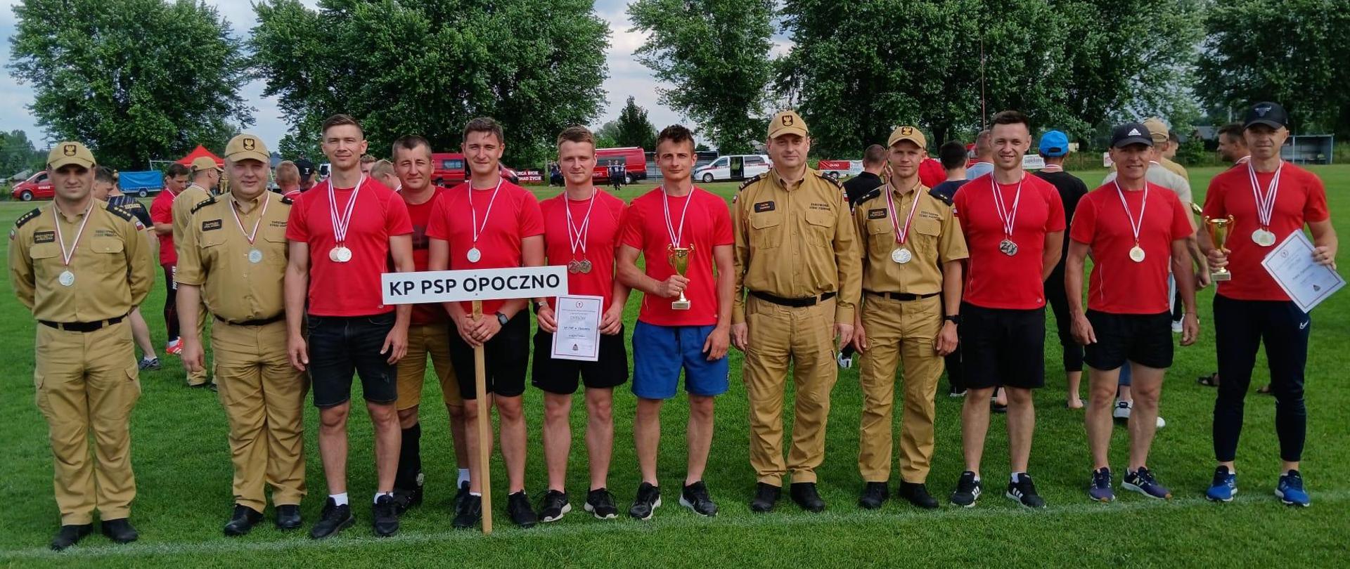 Na zdjęciu dwunastu funkcjonariuszy stających w jednym szeregu, czterech z nich w umundurowaniu służbowym, wszyscy mają zawieszone medale, trzech z nich trzyma puchary i dyplomy. Drużyna KP PSP Opoczno w czerwono czarnych strojach sportowych, jeden trzyma tablice z napisem KP PSP Opoczno. W tle inni funkcjonariusze i boisko sportowe