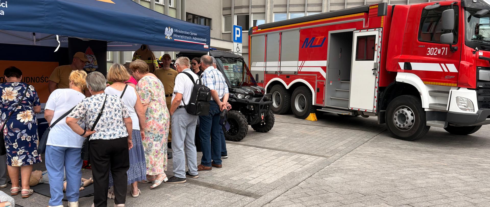 Fotografia przedstawia stoisko Komendy Miejskiej Państwowej Straży Pożarnej w Kielcach. W głębi stoi pojazd typu UTV oraz wóz gaśniczy a przy stoisku zebrali się zainteresowani uczestnicy festynu Akademii OLIOC.