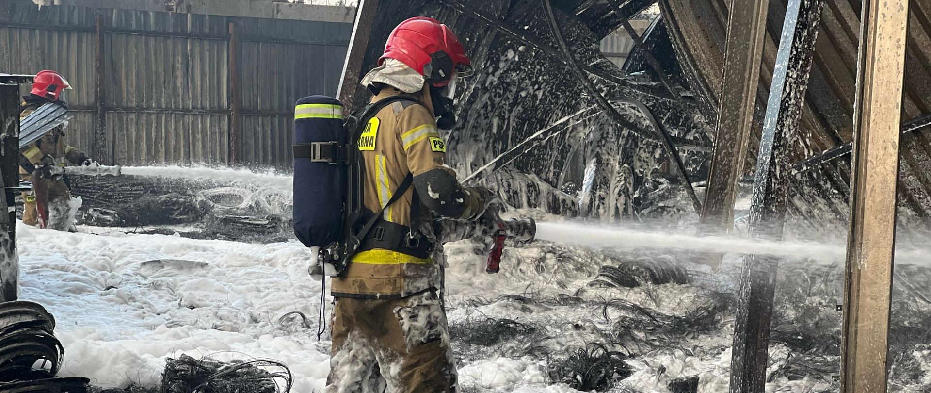 Zdjęcie przedstawia dwóch strażaków w akcji podczas gaszenia pożaru. Główny strażak w centralnej części kadru, ubrany w żółto-czarny kombinezon ochronny, czerwony hełm i maskę, trzyma wąż strażacki i kieruje strumień piany na płonącą konstrukcję. Drugi strażak znajduje się w tle po lewej stronie, również w pełnym wyposażeniu. Teren jest pokryty pianą gaśniczą i czarnym popiołem, a w tle widoczne są zniszczone elementy konstrukcji i płot. Sytuacja wygląda na intensywną walkę z ogniem w przemysłowym lub magazynowym obiekcie.
