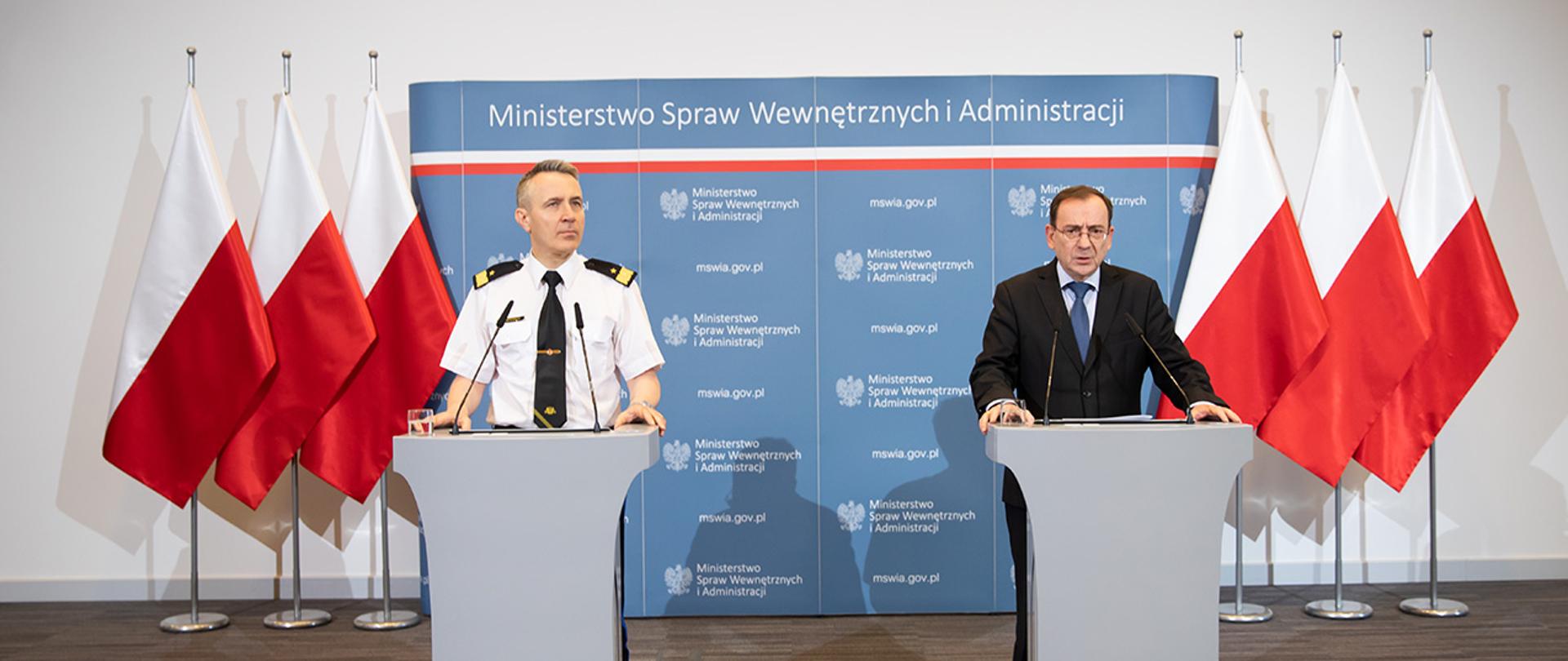 Na zdjęciu widać Mariusza Kamińskiego ministra SWiA i nadbryg. Andrzej Bartkowiak komendanta głównego Państwowej Straży Pożarnej stojących przy mównicach na tle ścianki MSWiA i flag Polski. Minister Kamiński przemawia.