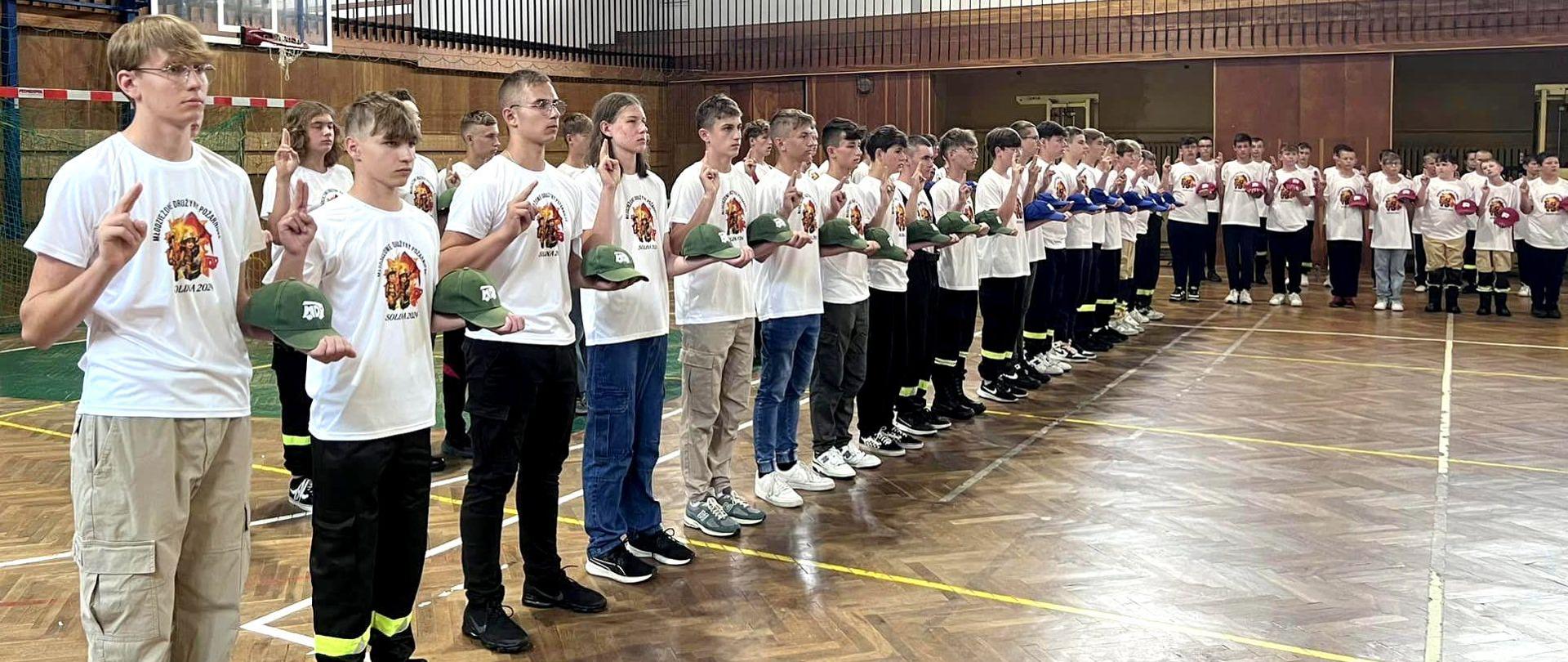 Zdjęcie zrobione w sali gimnastycznej podczas uroczystej zbiórki uczestników obozu Młodzieżowych Drużyn Pożarniczych. Uczestnicy obozu, członkowie Młodzieżowych Drużyn Pożarniczych składają ślubowanie.