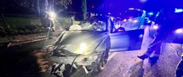 Zdjęcie przedstawia rozbite srebrne BMW w tle uszkodzone słupy energetyczne. Strażacy stoją przy samochodzie