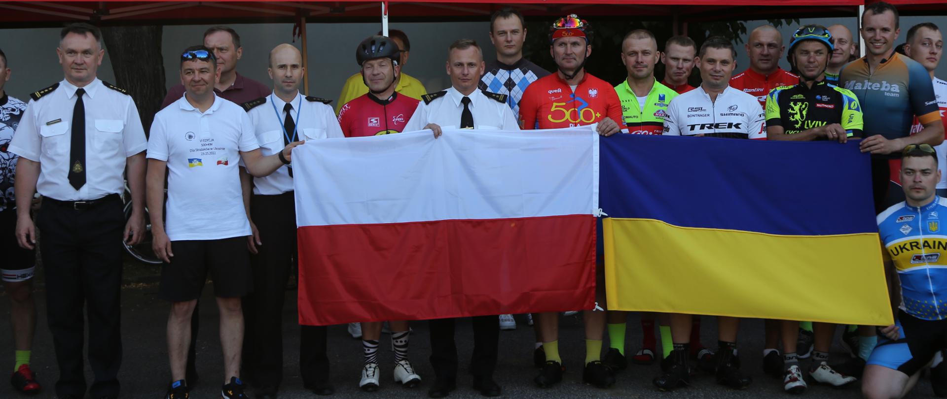 Grupowe zdjęcie uczestników maratonu kolarskiego w towarzystwie Zastępców Komendanta Centralnej Szkoły PSP (18 osób). Uczestnicy zdjęcia trzymają rozwinięte obok siebie flagi Polski i Ukrainy