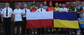 Grupowe zdjęcie uczestników maratonu kolarskiego w towarzystwie Zastępców Komendanta Centralnej Szkoły PSP (18 osób). Uczestnicy zdjęcia trzymają rozwinięte obok siebie flagi Polski i Ukrainy