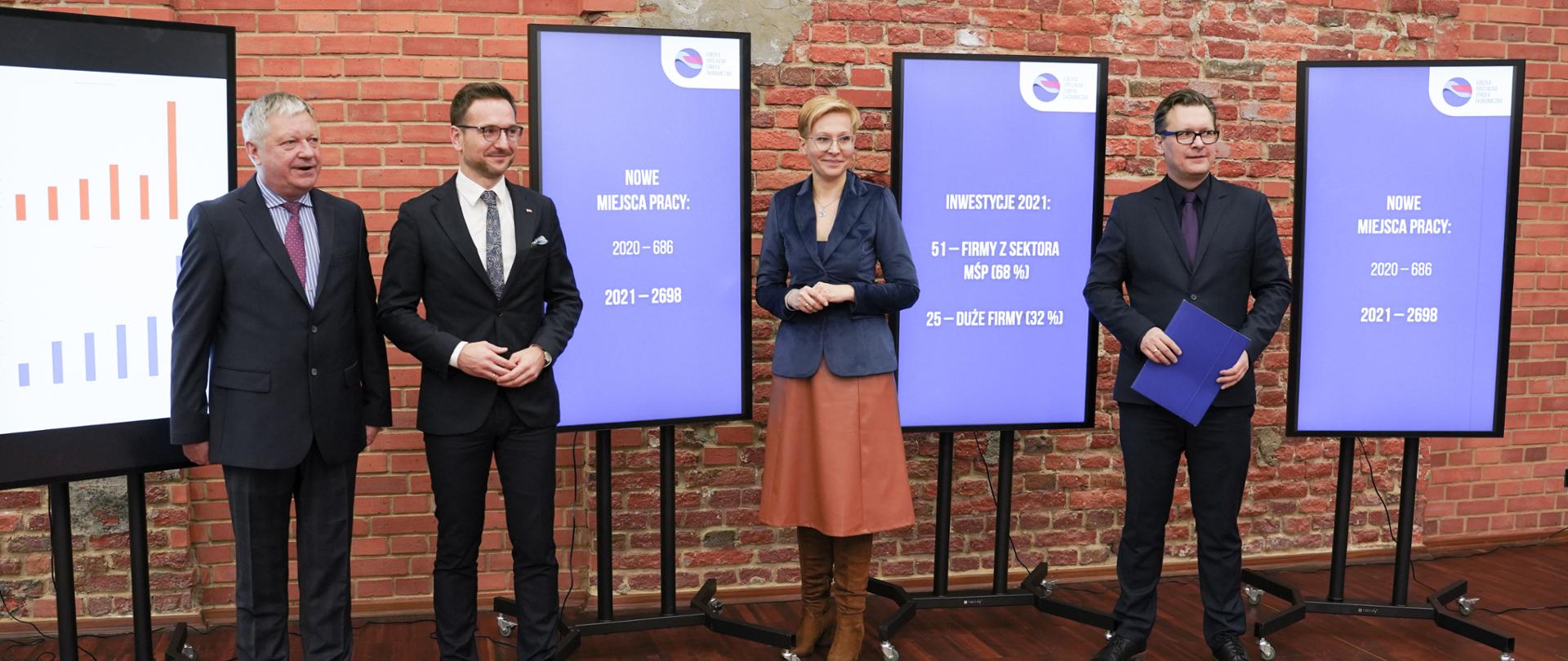 Na zdjęciu 4 osoby pomiędzy tablicami informacyjnymi o sukcesach Łódzkiej Specjalnej Strefy Ekonomicznej. Drugi od prawej minister Buda
