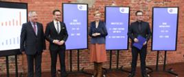 Na zdjęciu 4 osoby pomiędzy tablicami informacyjnymi o sukcesach Łódzkiej Specjalnej Strefy Ekonomicznej. Drugi od prawej minister Buda