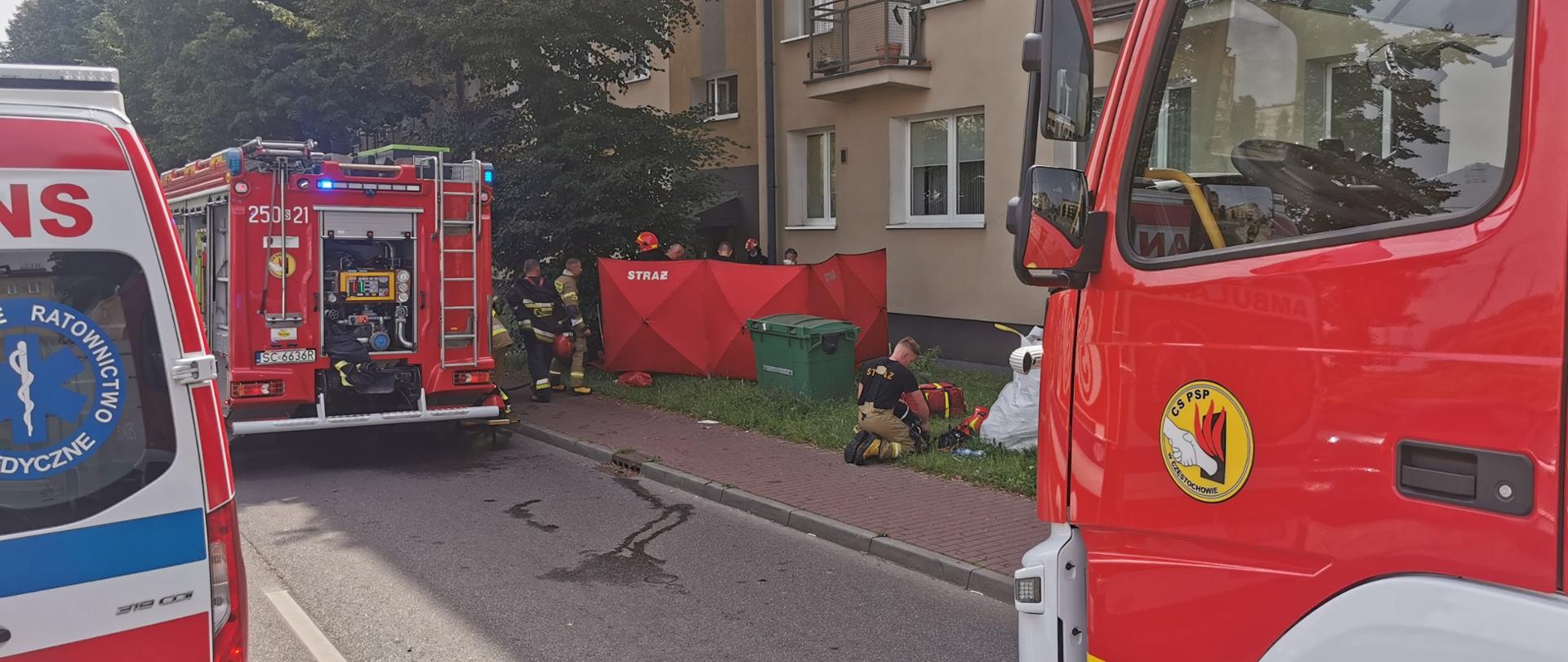 Częstochowa - pożar mieszkania - przed blokiem mieszkalnym samochód ratownictwa medycznego i 2 samochody pożarnicze, przy chodniku strażacy z rozłożonym parawanem